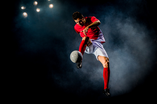 A soccer player wearing red forcefully kicks the ball into the air