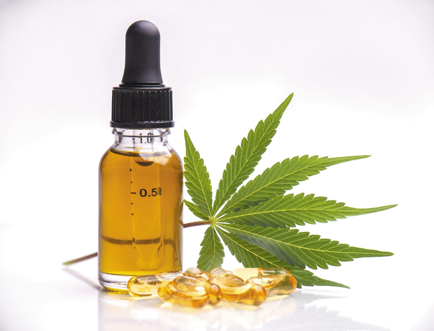 A closed transparent tincture bottle filled with CBD oil sitting beside a cannabis leaf and CBD capsules