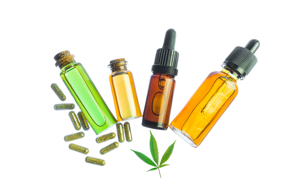 Two big and small closed transparent bottle filled with CBD oil laying close to a small closed brown tincture bottle filled with CBD oil and a big closed transparent bottle filled with CBD oil alongside few CBD capsules