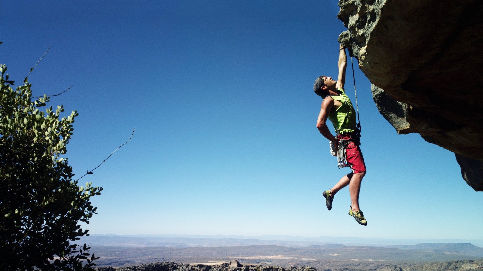 A man wearing a green t-shirt and red shorts with a rope tied around his waist is seen dangling with his left hand hanging on the side of the rock